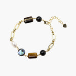 Gold chain bracelet with tiger's eye, obsidian, pearl, and iridescent abalone stones on cream background.