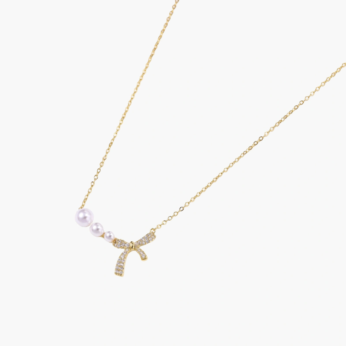 Delicate gold necklace with three white pearls and crystal-embellished bow pendant on cream background