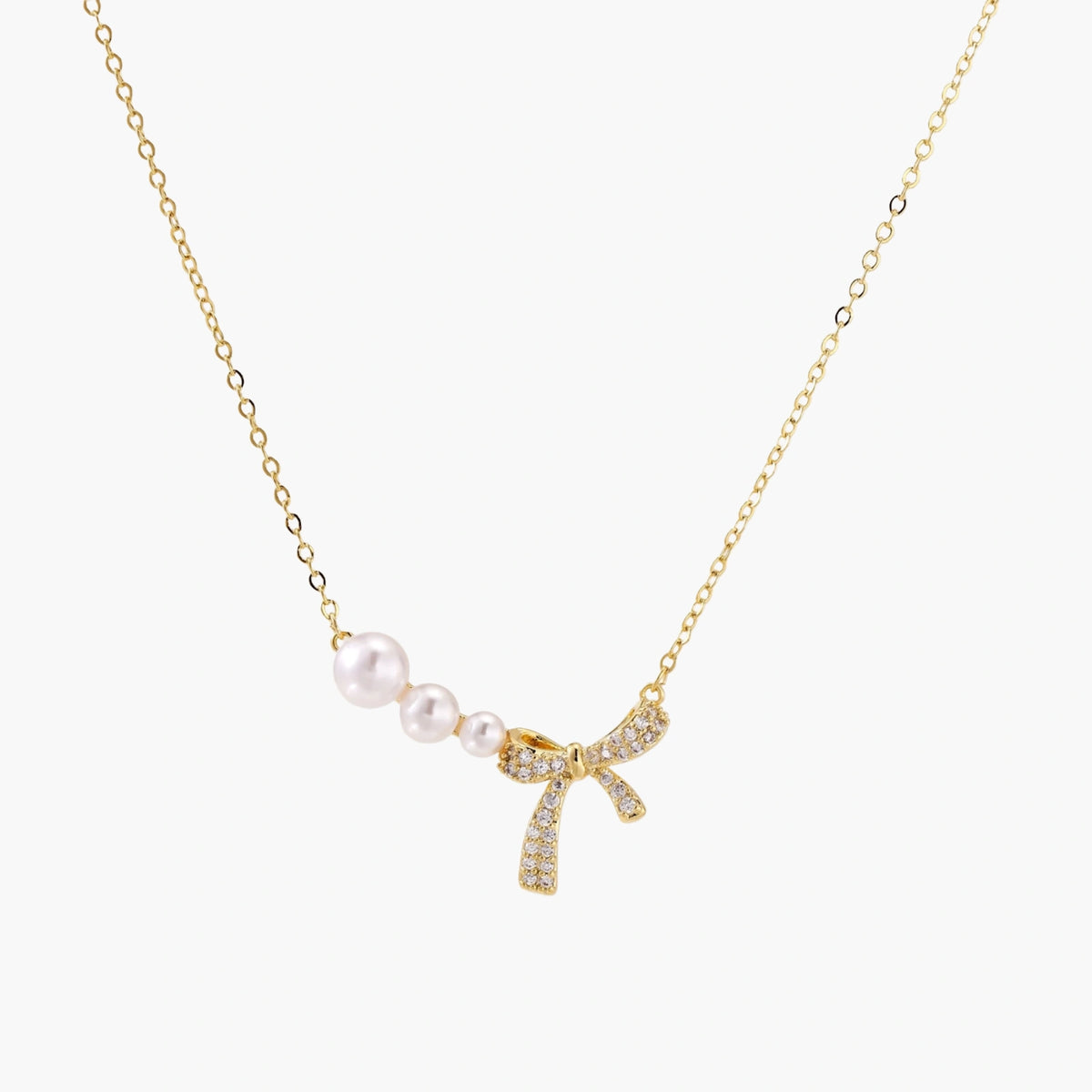 Delicate gold necklace with three white pearls and crystal-embellished bow pendant on cream background